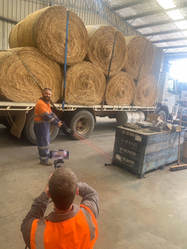 A worker in orange high-visibility clothing standing beside a truck loaded with round hay bales while someone films from behind.