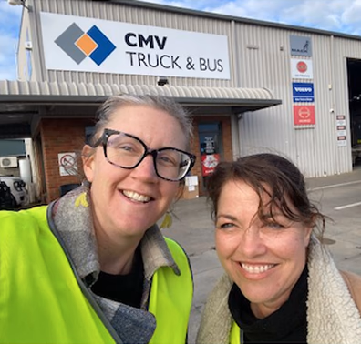 Two women in high-visibility vests smiling in front of a CMV Truck and Bus workshop.