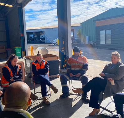 A small group seated in a circle at an open workshop doorway, most of them in orange high-visibility clothing.