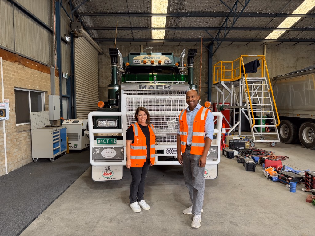 Two people in orange high-visibility vests standing in front of a green Mack truck inside a workshop.