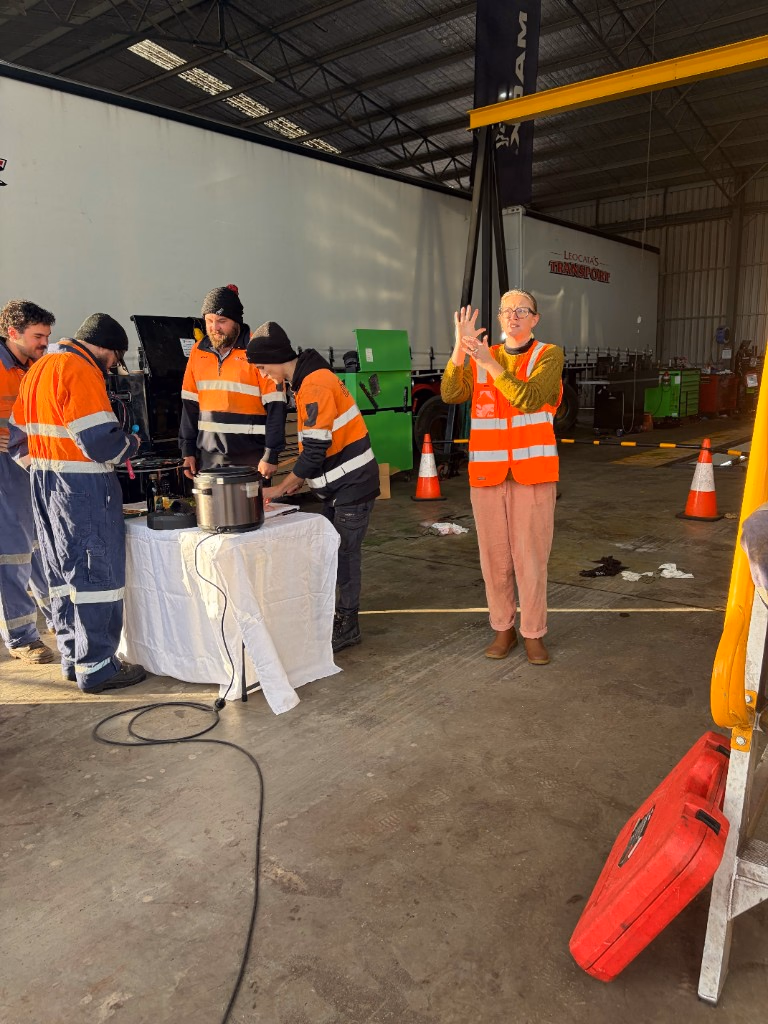 A woman in an orange high-visibility vest presenting with both hands raised beside a table holding a slow cooker, while workers in high-vis gather around.
