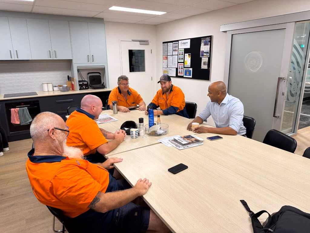 A team meeting around a timber table, with four people in orange high-visibility shirts and one person in a light blue shirt holding a coffee cup.